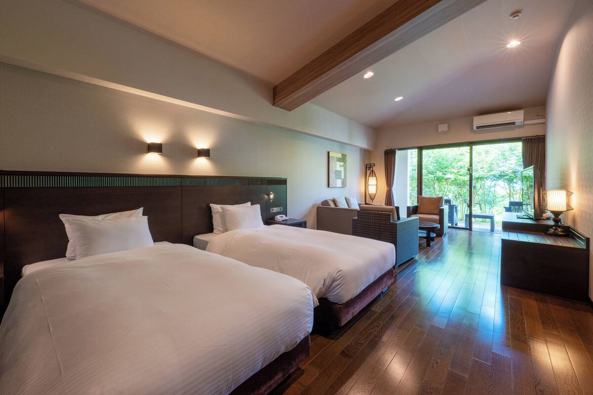 Une chambre d’hôtel avec deux lits, un coin salon avec une table et des chaises, et une grande fenêtre donnant sur l’extérieur.