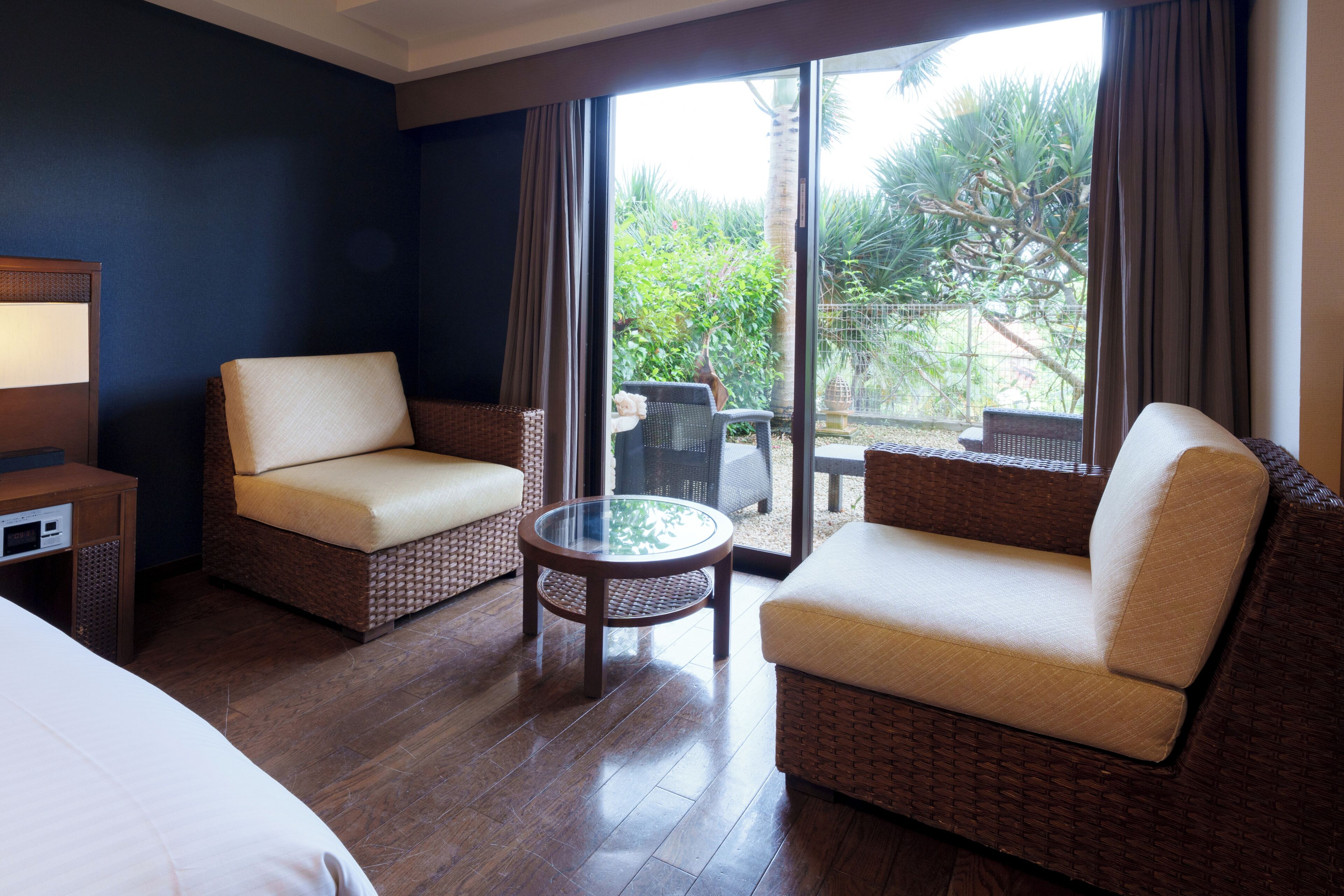 Habitación de hotel con una cama, dos sofás de mimbre, una mesita redonda y un amplio ventanal corredero que da a un área exterior con vegetación.