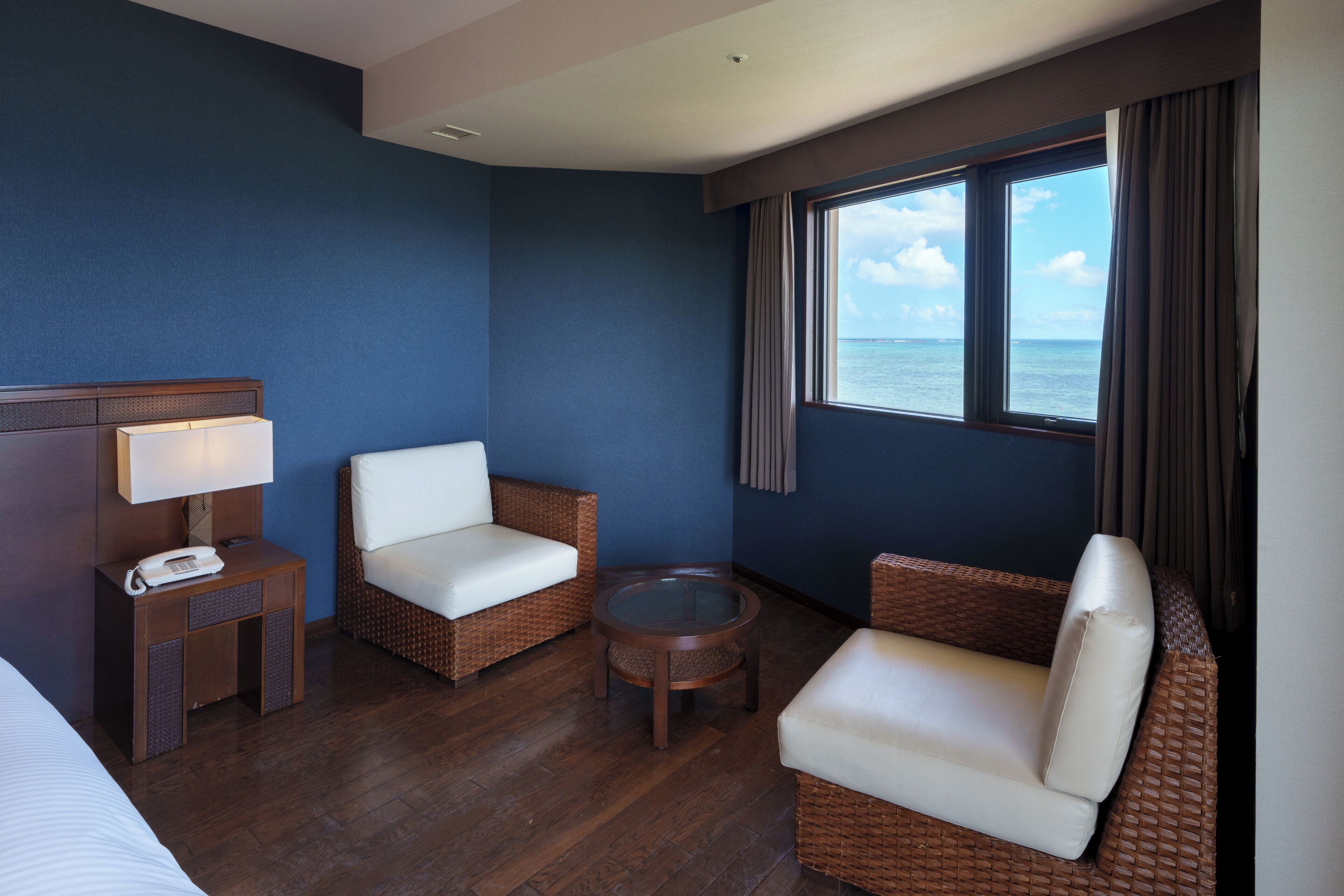 A hotel room with a bed, two wicker chairs, a small table, a lamp, and a window with a view of the sea.
