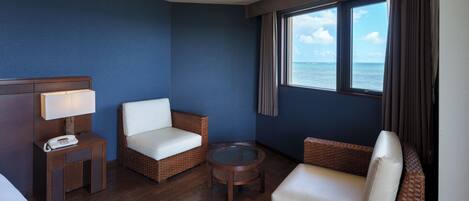 A hotel room with a bed, two wicker chairs, a small table, a lamp, and a window with a view of the sea.