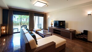 A modern hotel room with a large sofa, a flat-screen TV, a coffee table, and a sliding glass door leading to a balcony with a view of palm trees. - Grandvrio Resort Ishigakijima (Ishigaki)