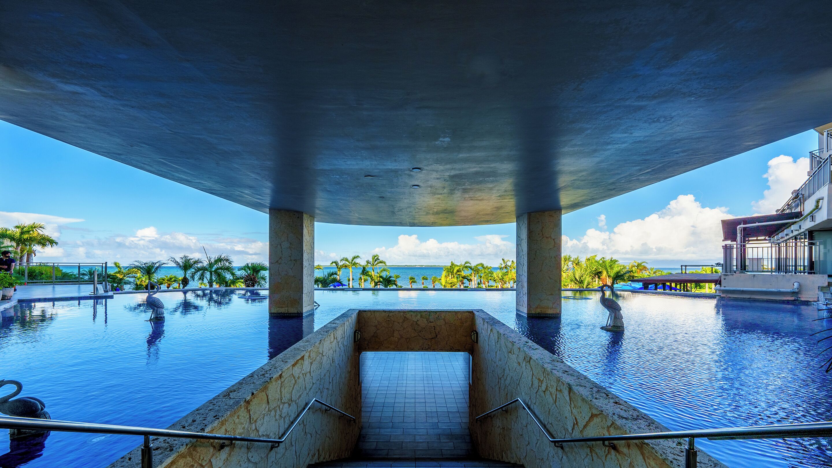 Ein Schwimmbad mit Meerblick, eine Treppe, die zu einer höheren Ebene führt, und ein Gebäude mit Balkonen.