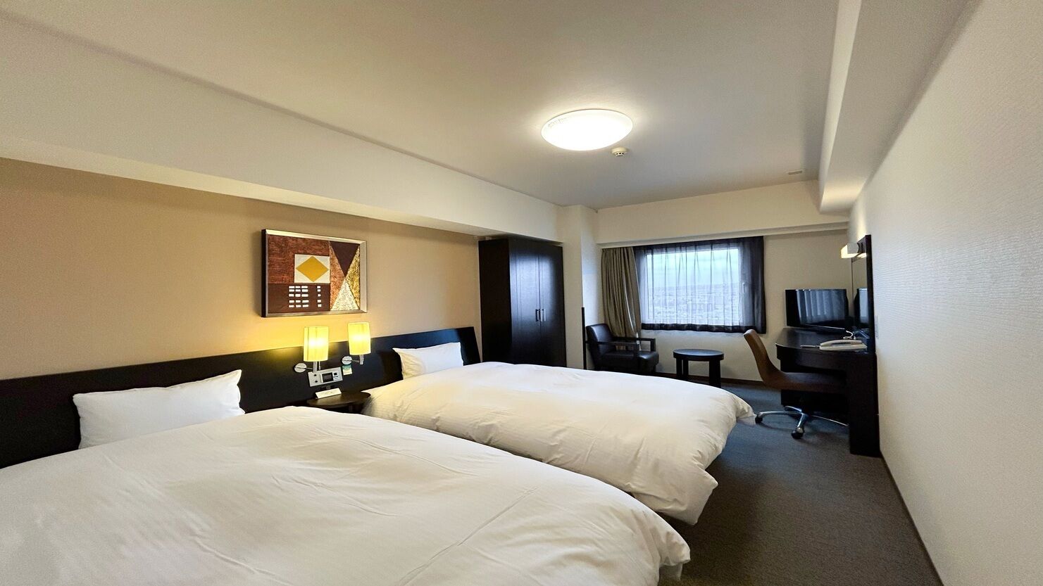 A hotel room with two beds, a desk, a chair, a TV, and a window with curtains.