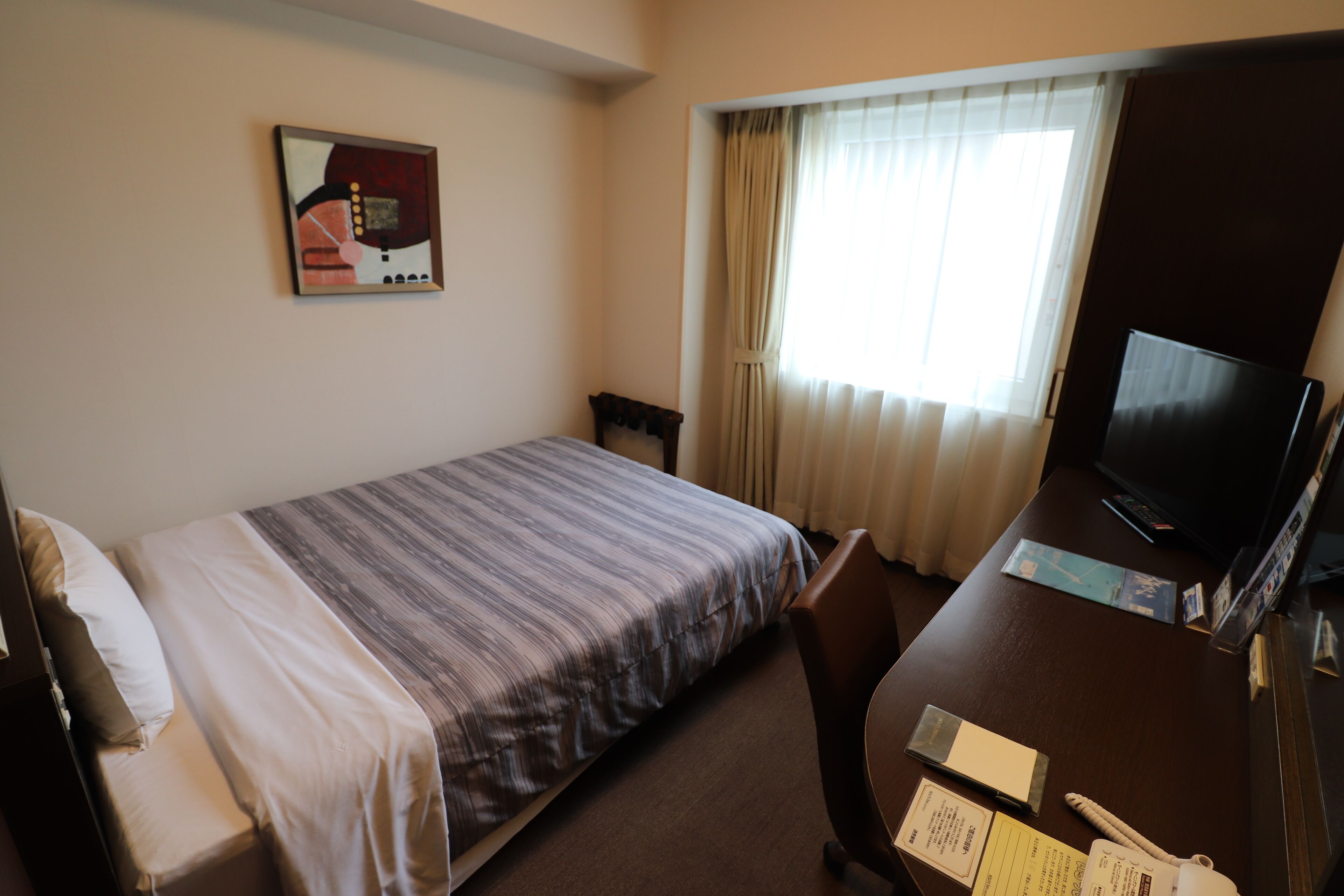 A hotel room with a bed, a desk with a television, a chair, and a window with curtains.
