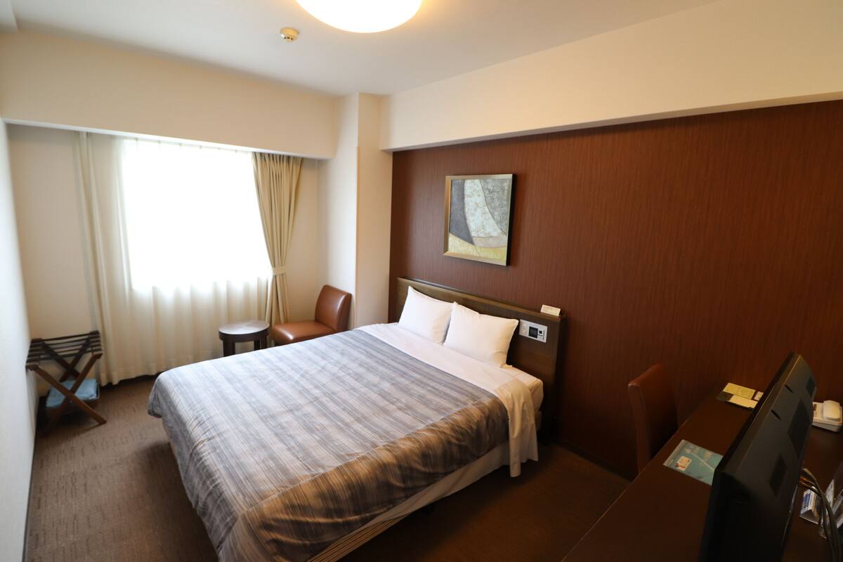 A hotel room with a large bed, a desk, a chair, a window with curtains, and a painting on the wall.