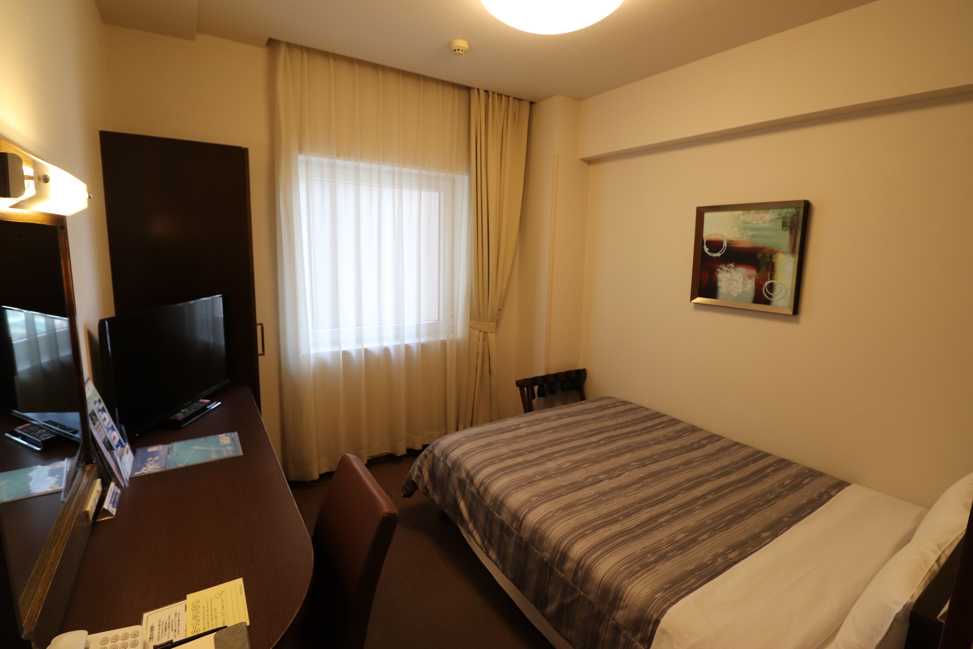 Habitació d'hotel amb un llit, una taula de treball amb televisió, una cadira i una finestra amb cortines.