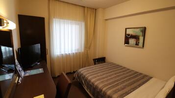 Habitació d'hotel amb un llit, una taula de treball amb televisió, una cadira i una finestra amb cortines.