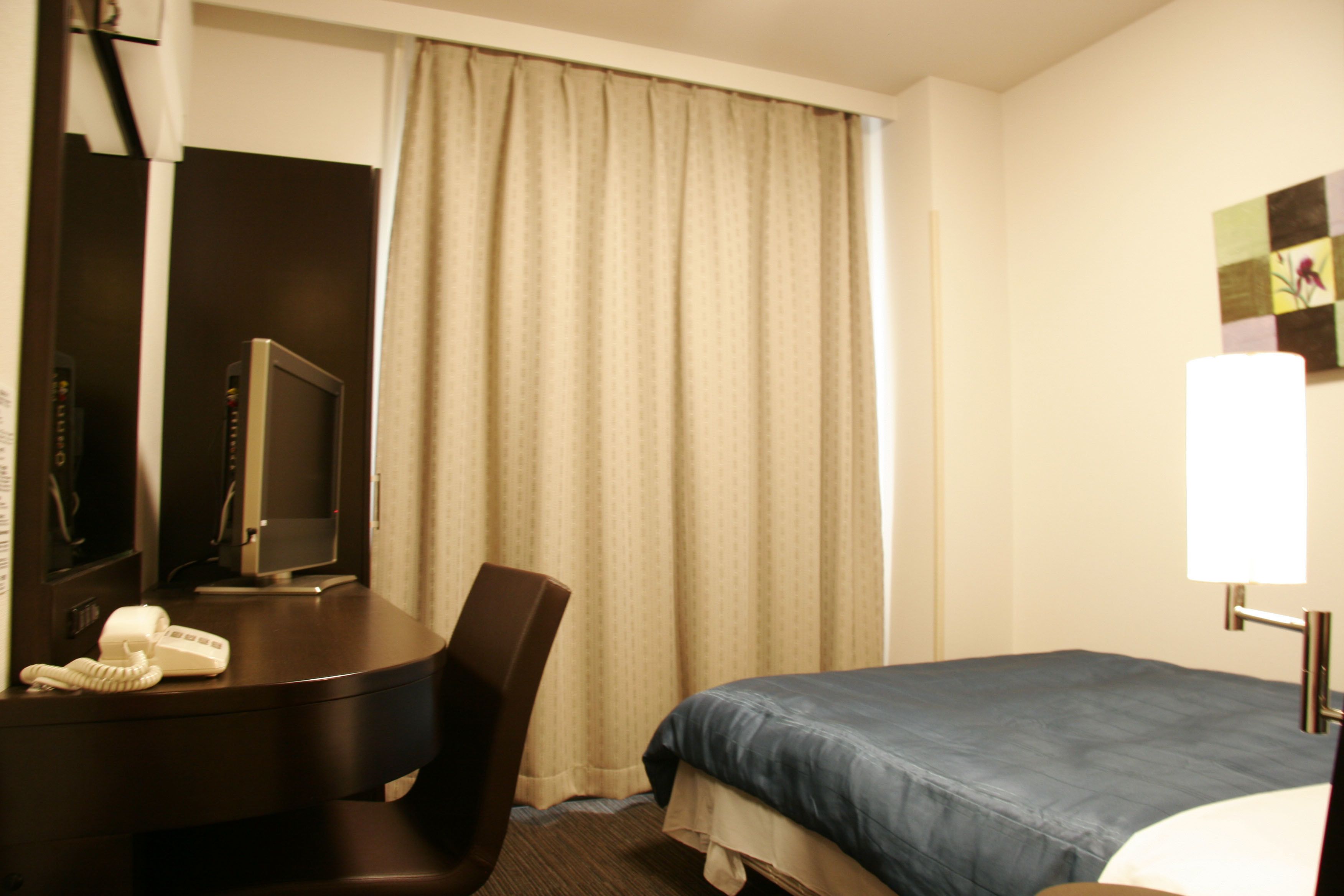 A hotel room with a bed, a desk with a telephone, a chair, a television, and a window with curtains.