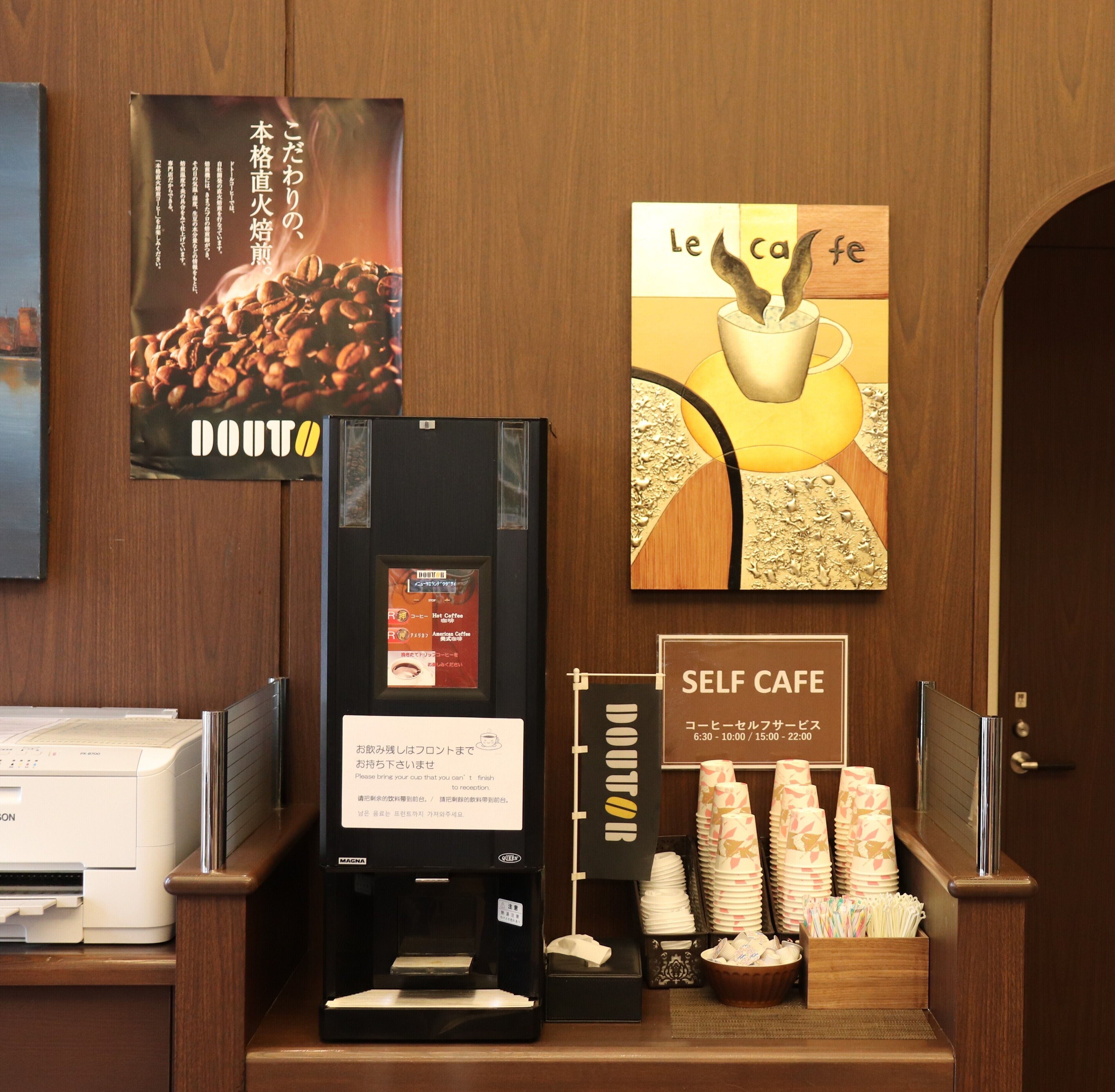 A self-service coffee station with a coffee machine, cups, and a sign indicating operating hours.