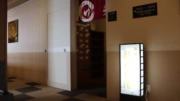 A hotel hallway with a checkered floor, a closed door, a wall-mounted sign with room numbers, and a digital display with Japanese text.