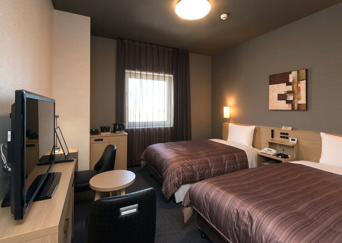 Hotelkamer met twee bedden, een bureau met computer, een stoel, een kleine tafel en een raam met gordijnen.