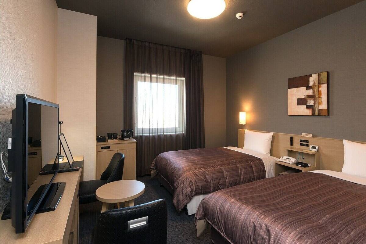 A hotel room with two beds, a desk with a computer, a chair, a small table, and a window with curtains.