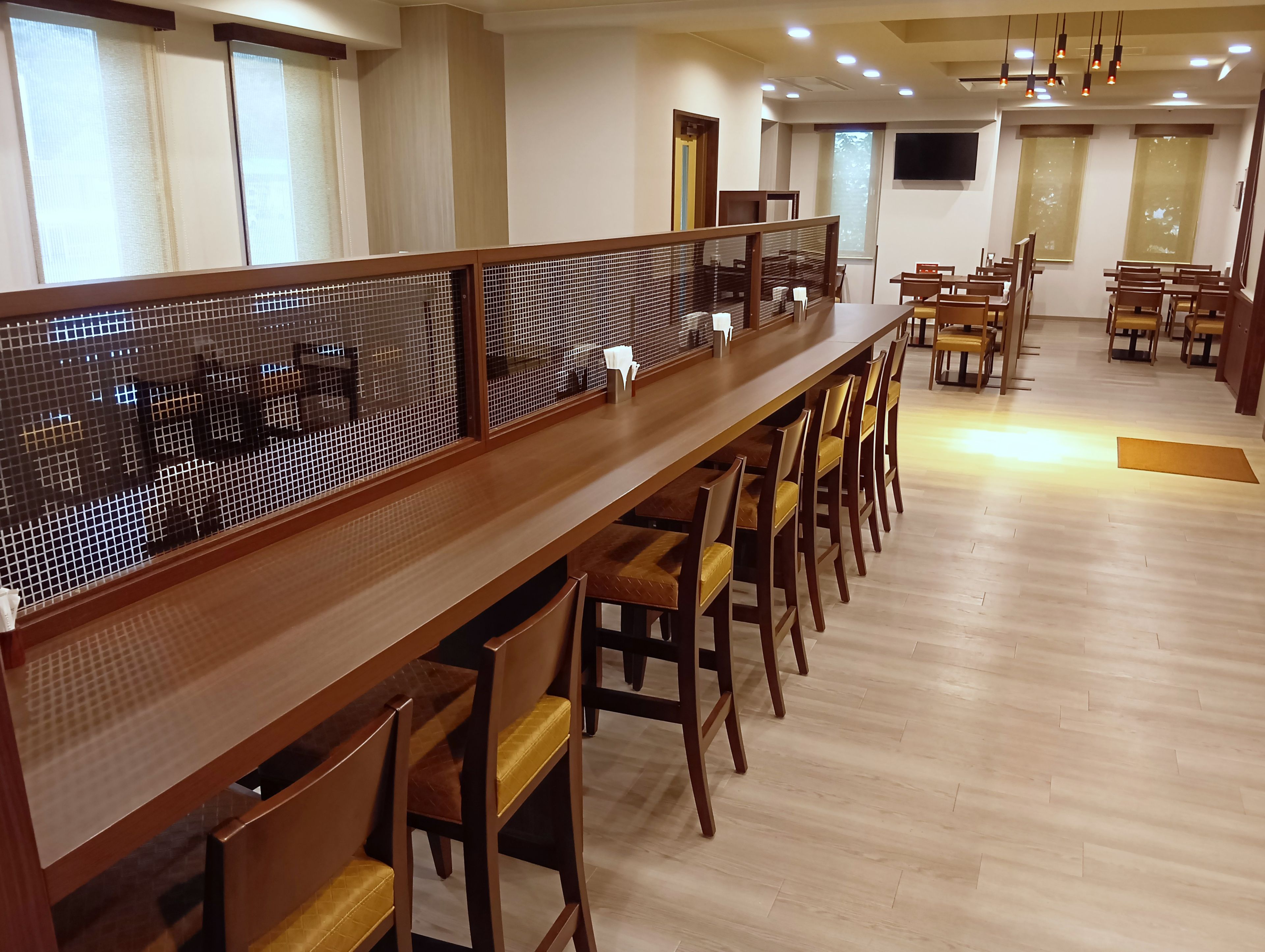 Una larga mesa de comedor con sillas, separada por una mampara de madera, en una habitación con suelo de madera y un televisor de pantalla plana montado en la pared.