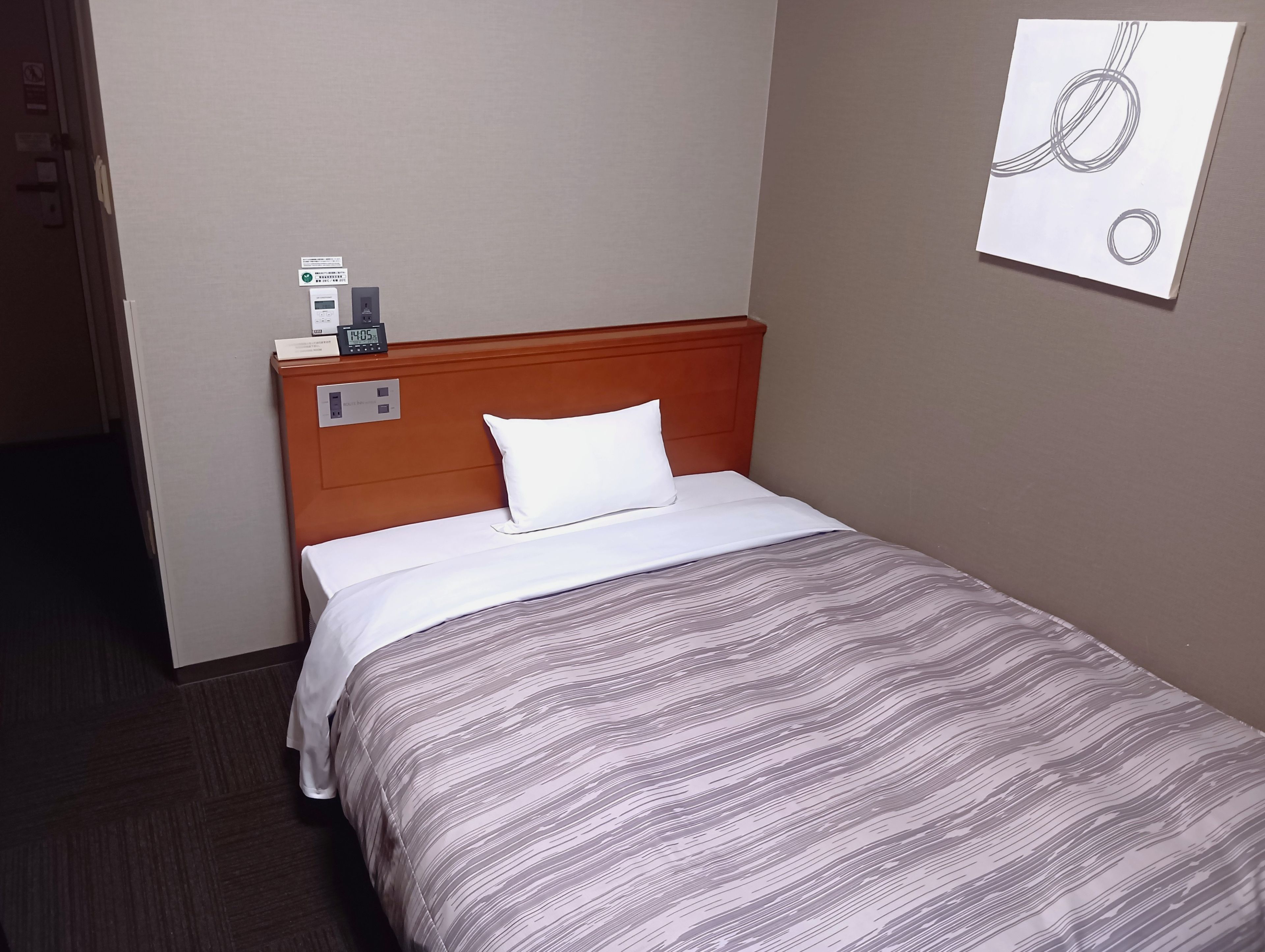 Habitación de hotel con cabecera de madera, almohada blanca, colcha a rayas, un reloj y una obra de arte colgada en la pared.