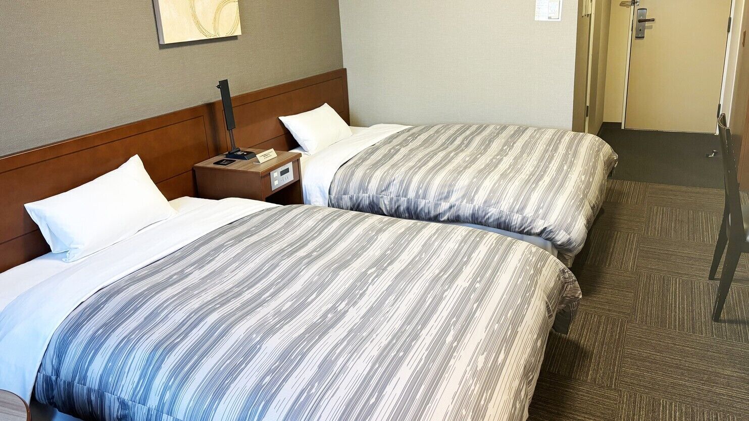 A hotel room with two beds, a nightstand, a lamp, and a door.
