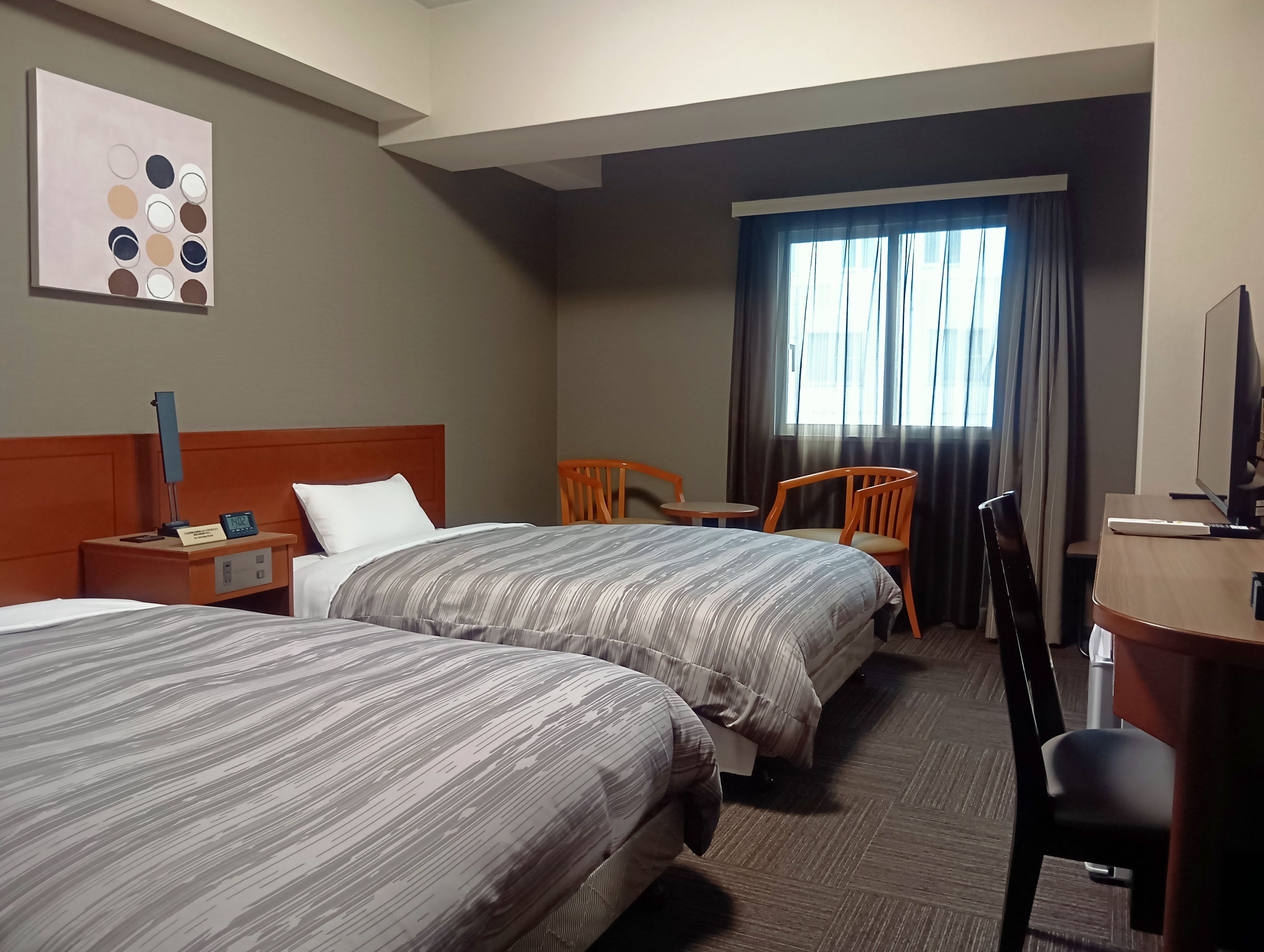 Ein Hotelzimmer mit zwei Betten, einem Schreibtisch, einem Stuhl, einem Fernseher und einem Fenster mit Vorhängen.