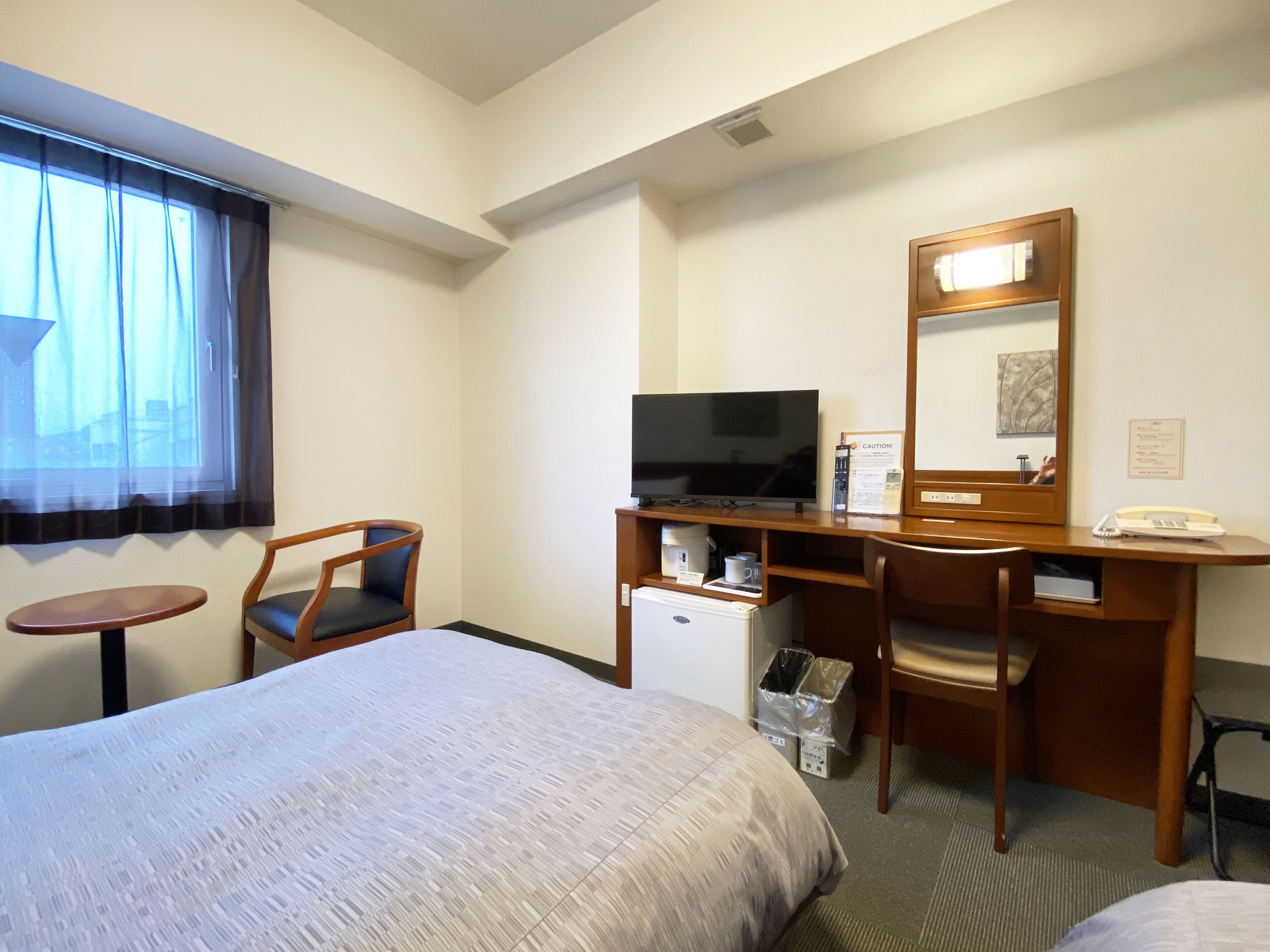 Habitación de hotel con cama, escritorio con computadora, televisión, refrigerador pequeño, silla, mesa y ventana con cortinas.