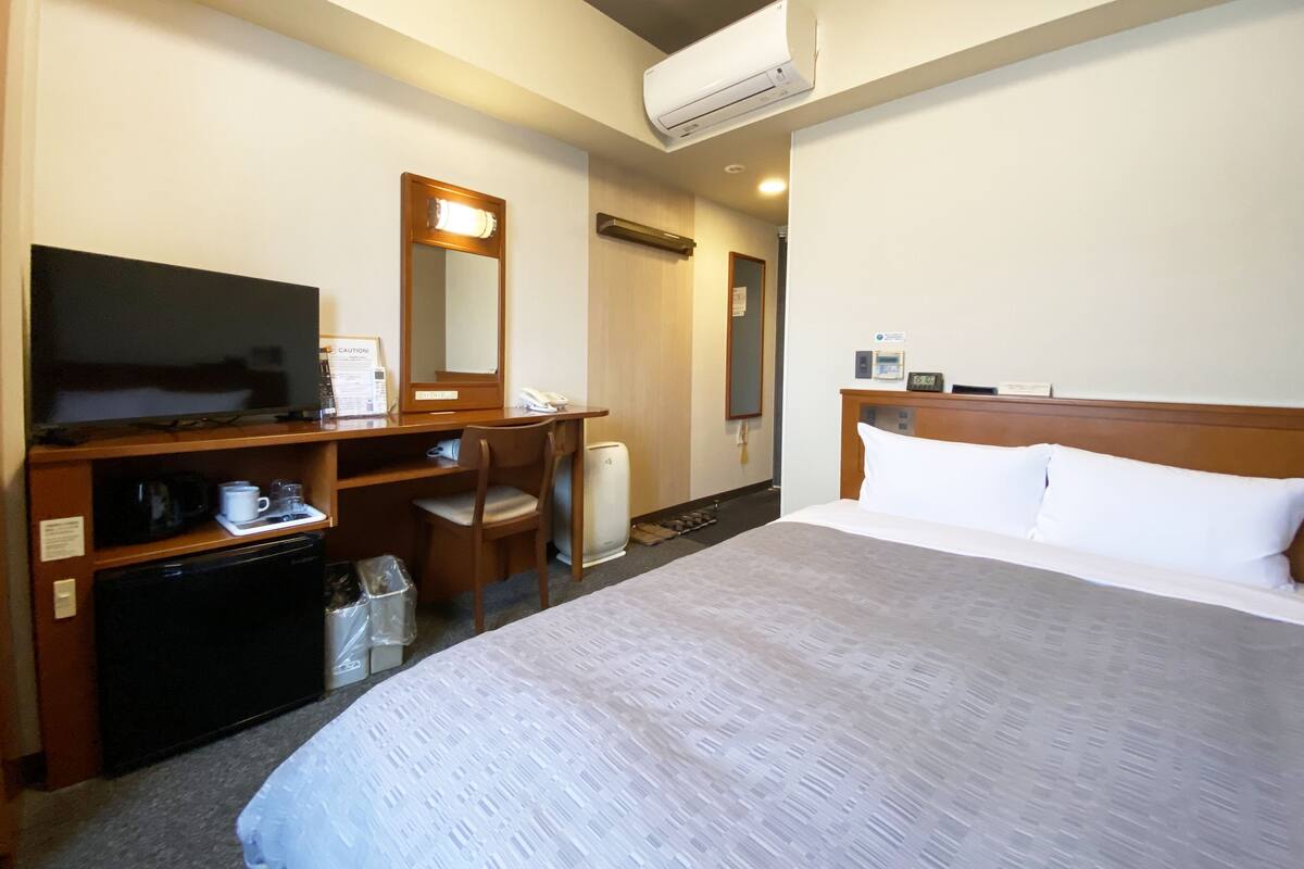 A hotel room with a bed, a desk with a television, a chair, a mirror, and an air conditioning unit.