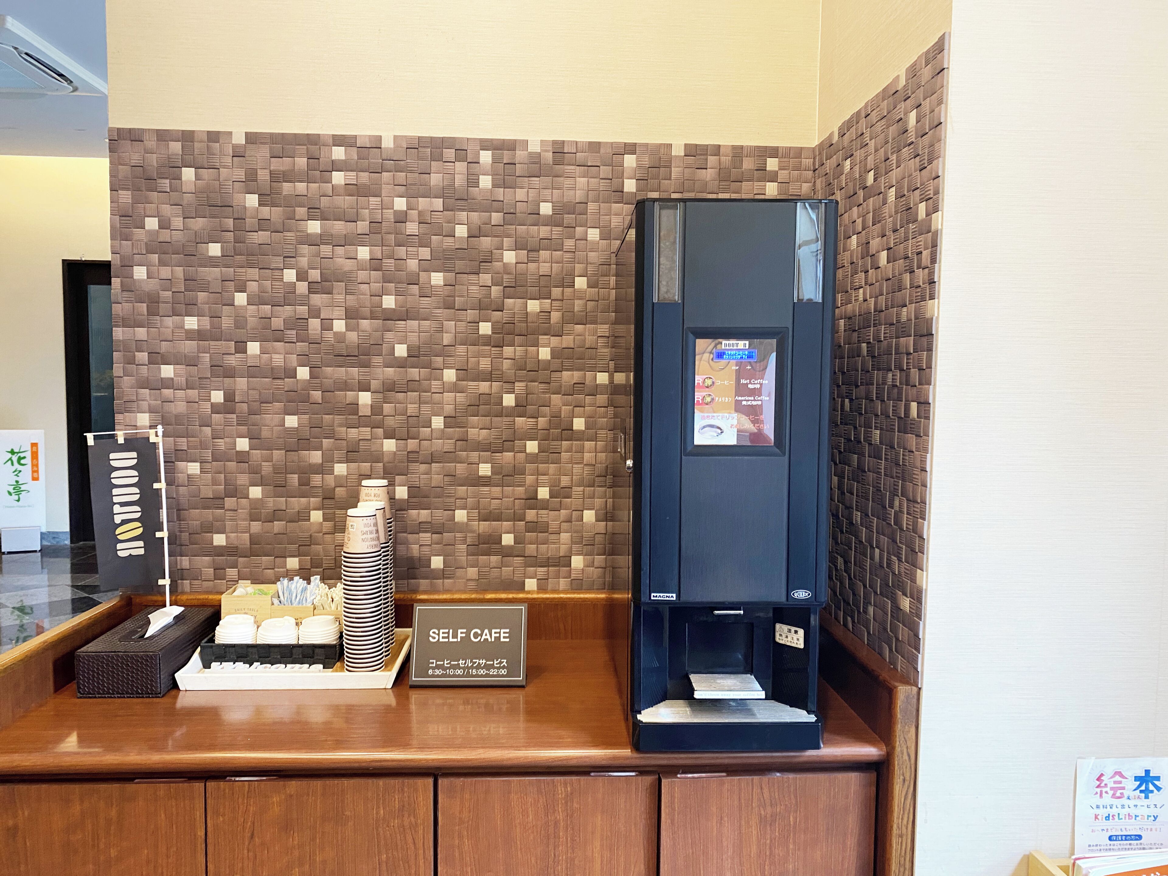 A self-service coffee station with a digital menu display, a stack of disposable cups, and a tissue box on a wooden counter.