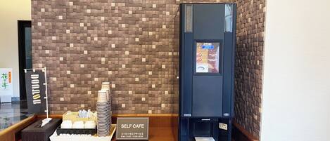 A self-service coffee station with a digital menu display, a stack of disposable cups, and a tissue box on a wooden counter.