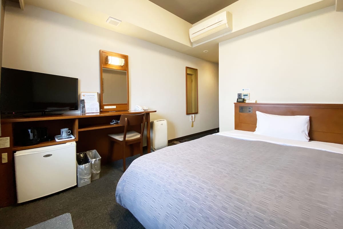 A hotel room with a bed, a desk with a television, a mirror, and a small refrigerator.