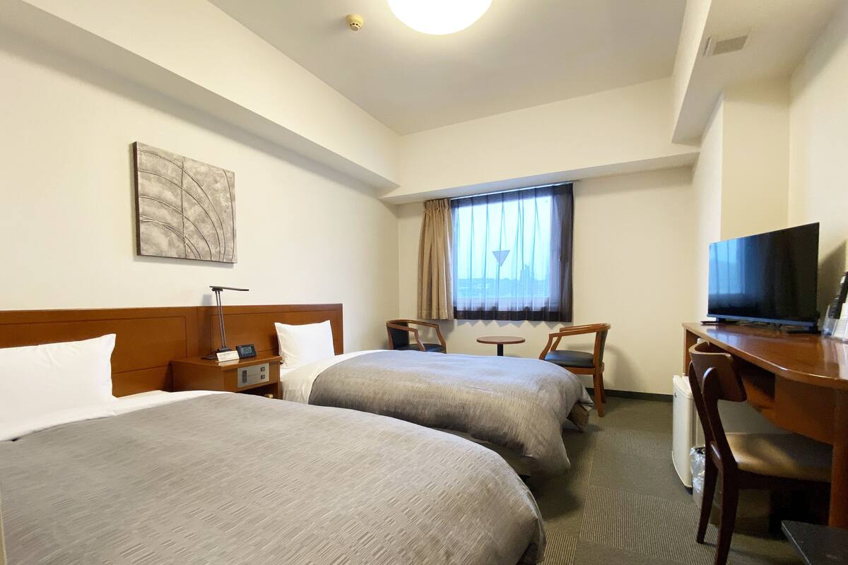 A hotel room with two beds, a desk with a television, a small table, and a window with curtains.