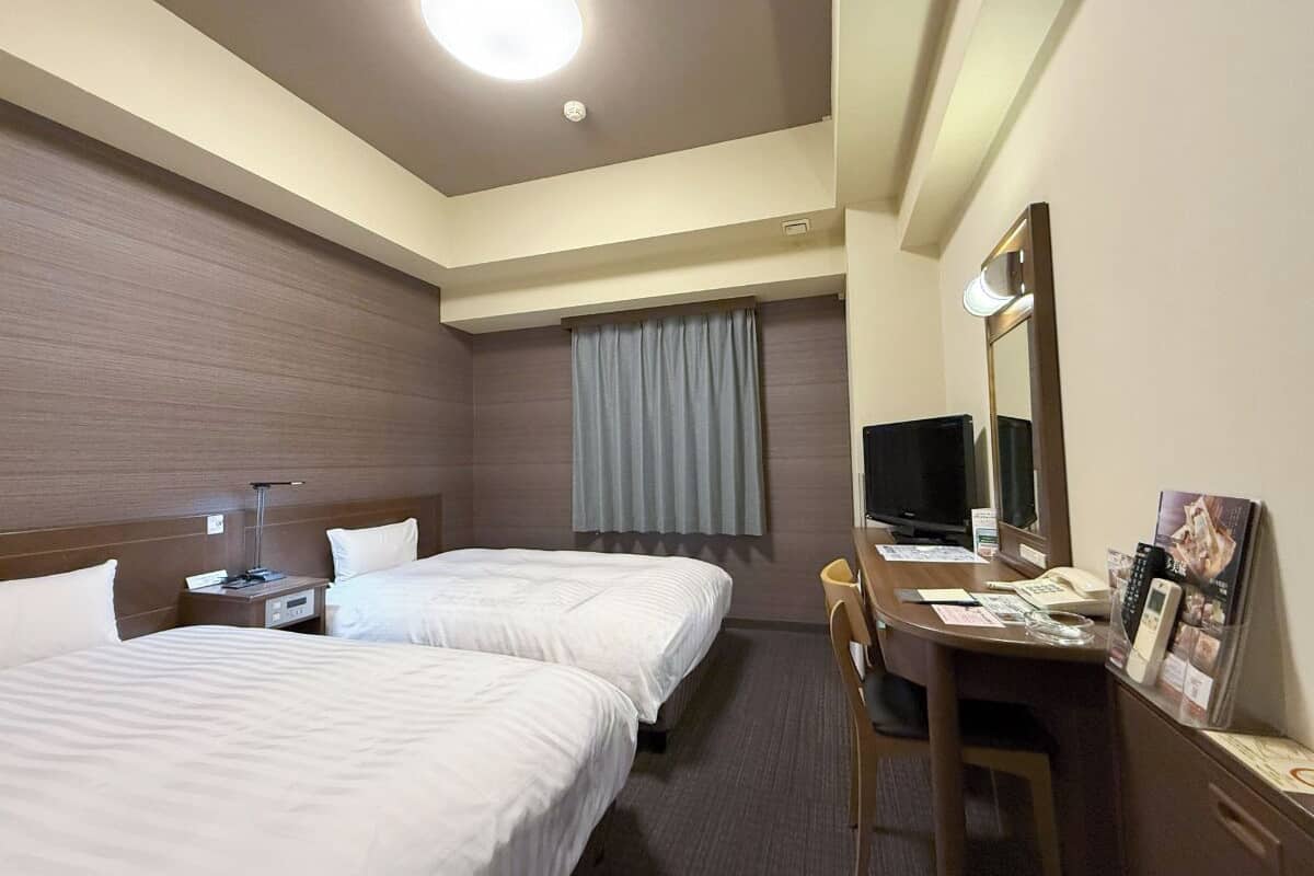 A hotel room with two beds, a desk with a chair, a television, a mirror, and a window with curtains.
