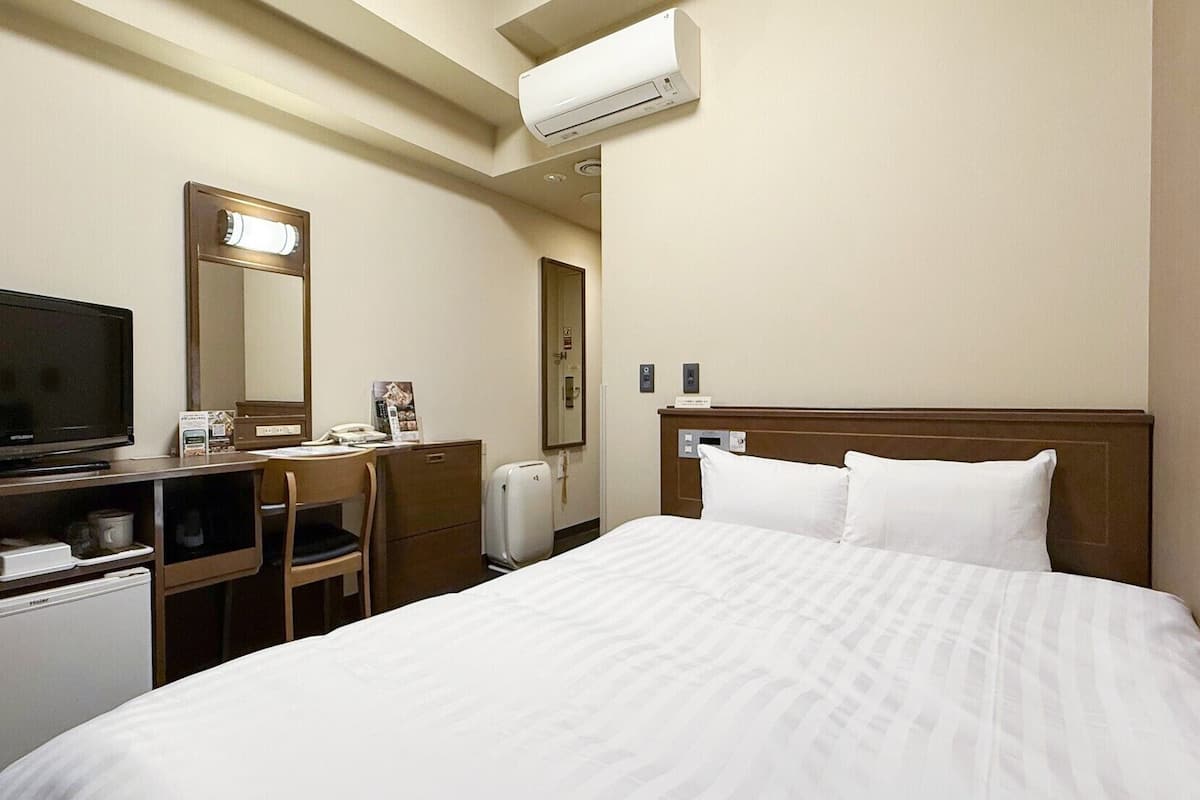 A hotel room with a bed, a television, a desk with a computer, a mirror, and an air conditioning unit.