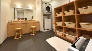 A bathroom with a wooden vanity, two stools, a washing machine, and wicker baskets on shelves. - Hotel Route Inn Hakata Ekimae (Fukuoka)