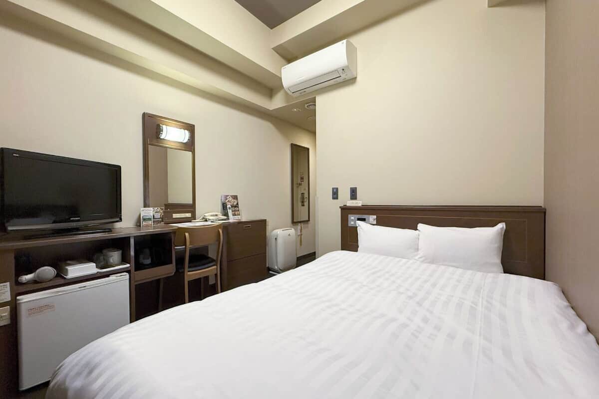 A hotel room with a bed, a desk with a television, a mini-fridge, a suitcase, and a mirror.