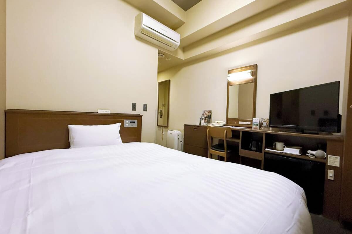 A hotel room with a large bed, a desk with a chair, a television, and an air conditioning unit.