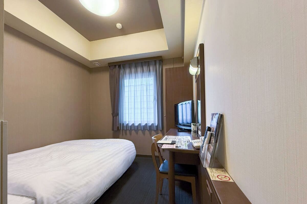 A hotel room with a single bed, a desk with a computer, a television, and a window with curtains.