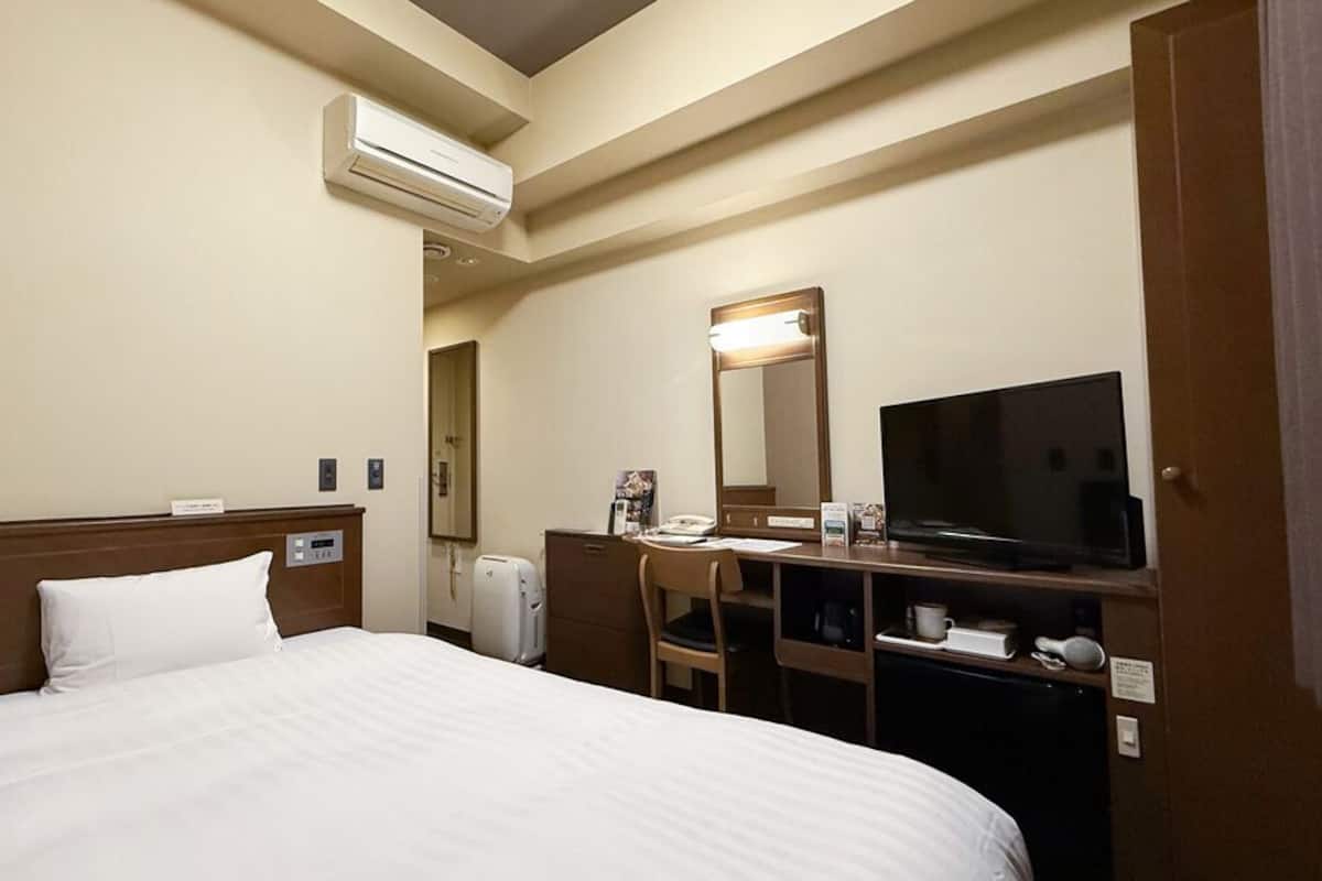 A hotel room with a bed, a desk, a television, a mirror, and an air conditioning unit.