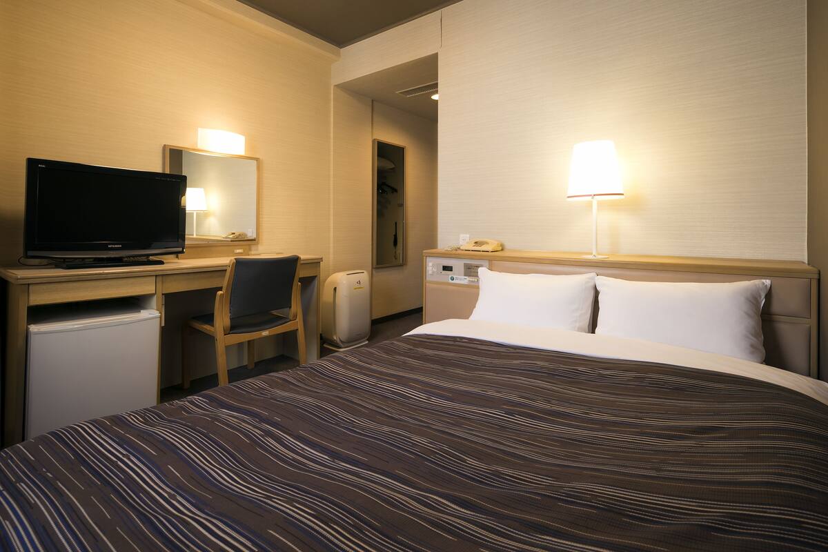 Habitació d'hotel amb un llit, una taula de treball amb televisió, una cadira, un nevera petit, una làmpada de nit i un telèfon.