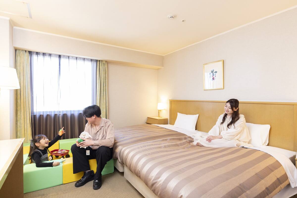 Habitación de hotel con cama, escritorio, ventana con cortinas y un niño jugando con un coche de juguete.