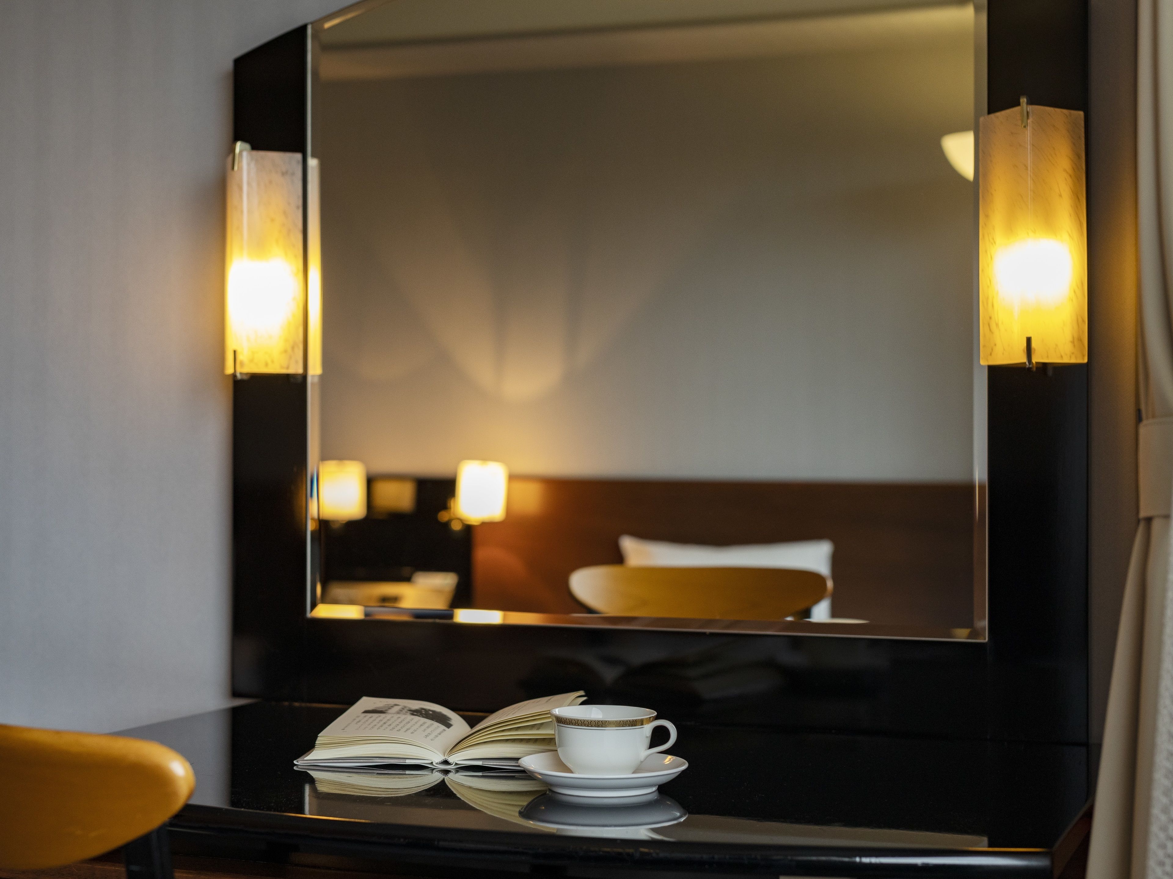 Camera d'albergo con un ampio specchio che riflette un letto, un comodino, un libro e una tazza su un tavolo.