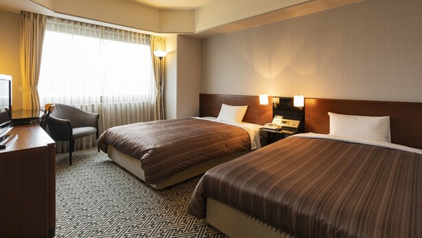 Habitación de hotel con dos camas, un escritorio con silla, una lámpara y una ventana con cortinas.