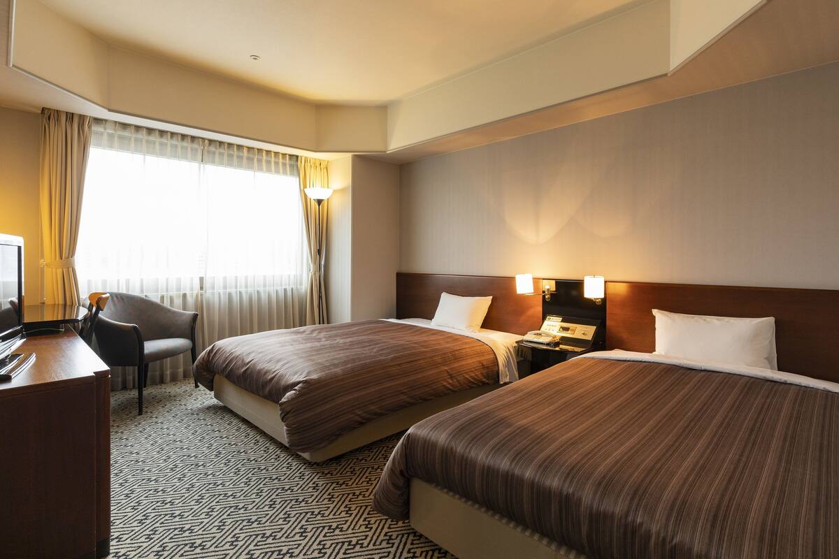 A hotel room with two beds, a desk with a chair, a lamp, and a window with curtains.