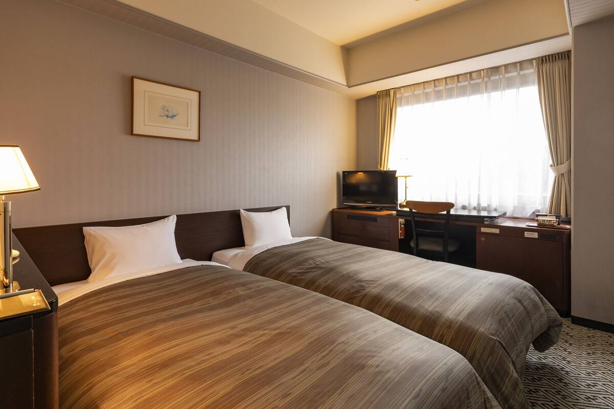A hotel room with two beds, a TV, a desk, and a window with curtains.