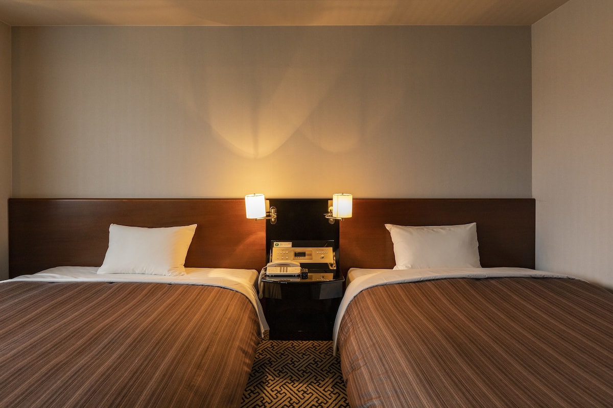 A hotel room with two beds, a nightstand with a phone, and wall-mounted lamps.