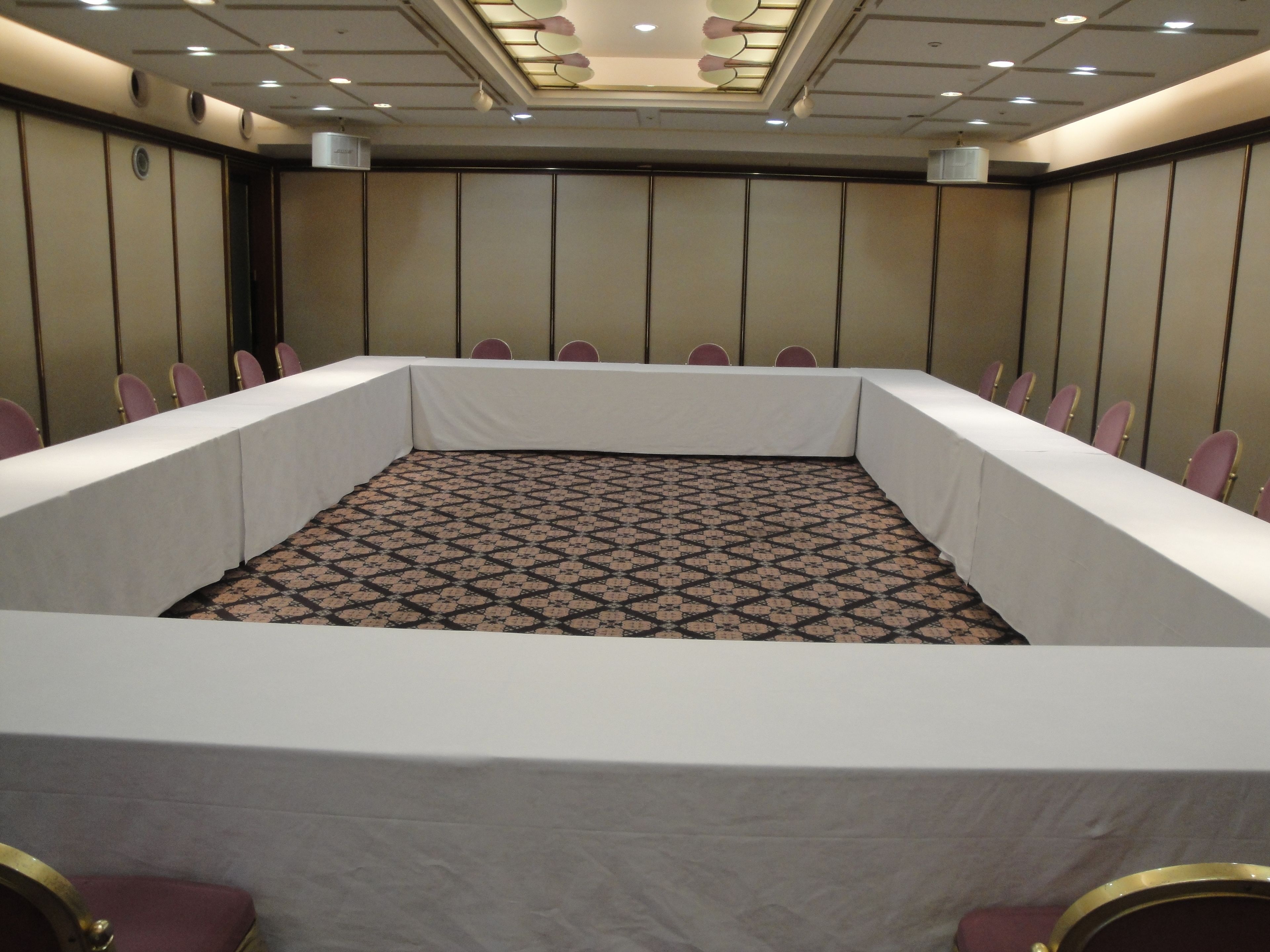 Sala de conferência com uma mesa comprida, cadeiras, carpete estampado e teto com iluminação embutida.