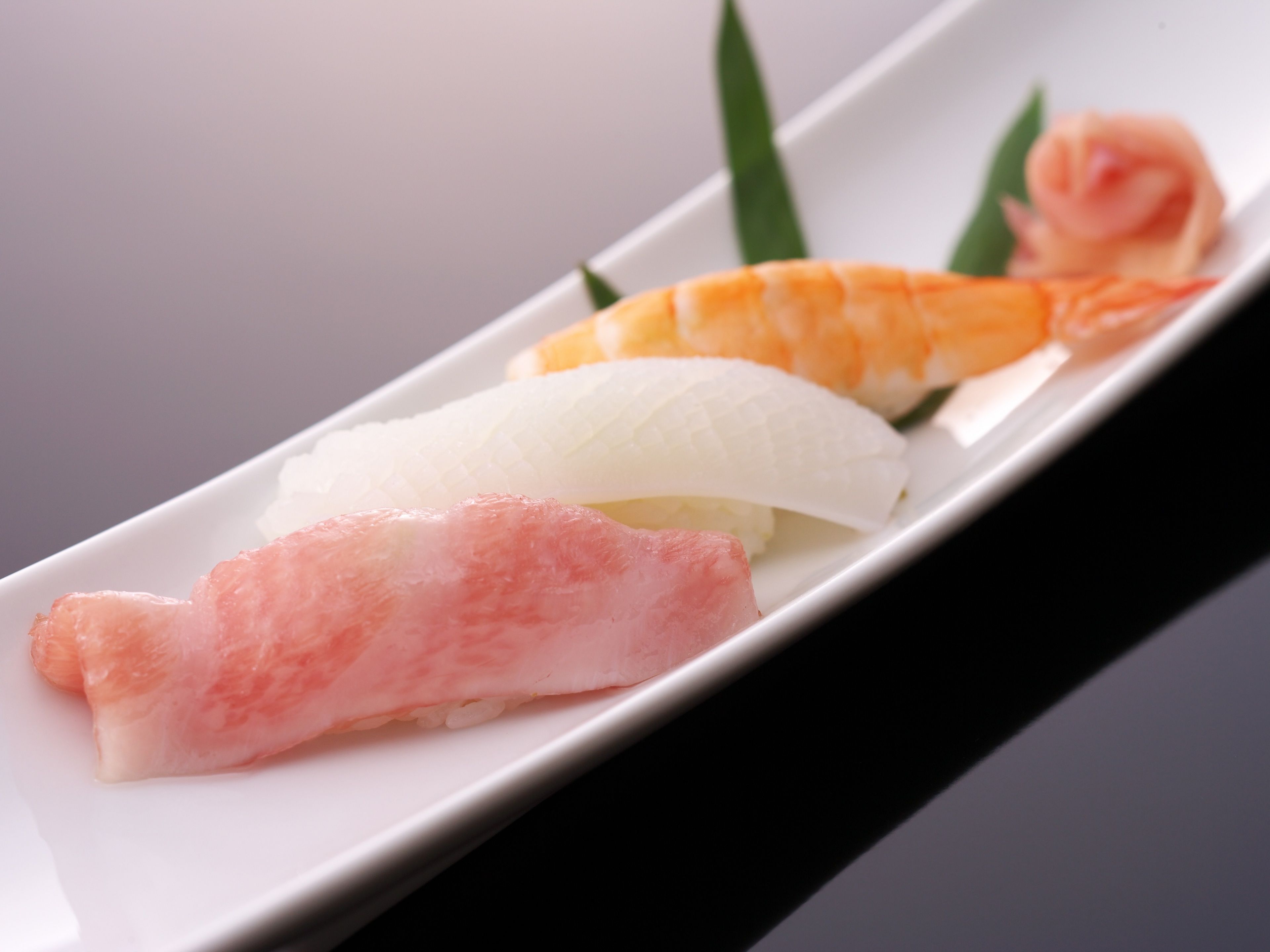 Un plato de sushi con tres piezas, incluyendo salmón, atún y pescado blanco, sobre un plato blanco adornado con una hoja verde.