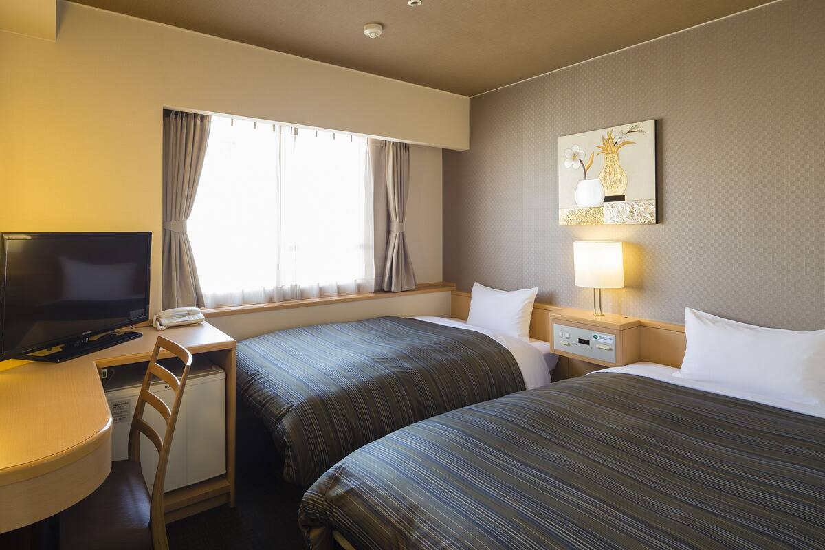 Une chambre d’hôtel avec deux lits, un bureau avec une télévision, une chaise, une table de chevet avec une lampe et une fenêtre avec des rideaux.