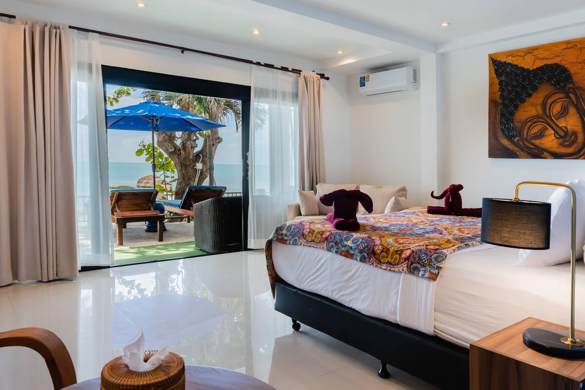 A bedroom with a bed, a nightstand, a lamp, a window with curtains, and a view of the beach.