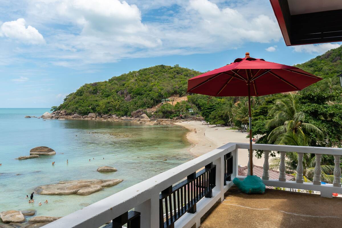 A beach view from a balcony with a red umbrella, clear turquoise water, and a rocky shoreline.