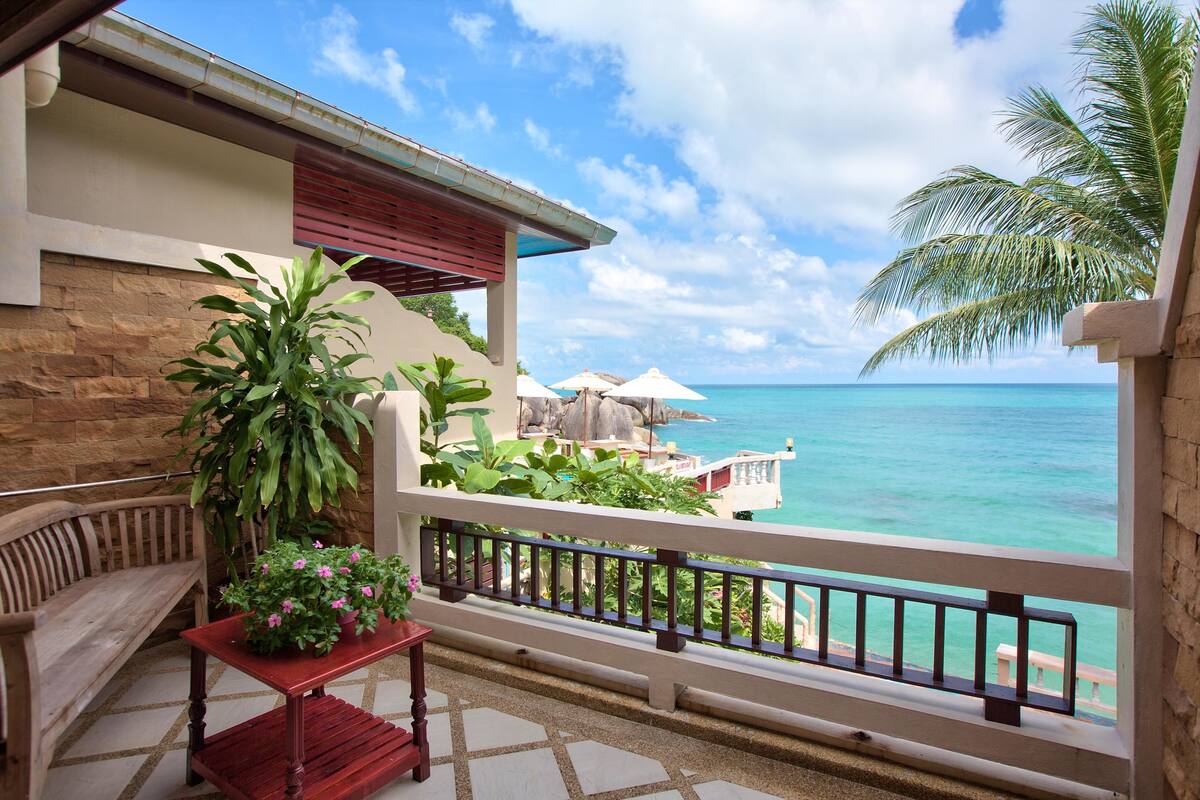 A balcony with a view of the ocean, a wooden bench, a table with potted plants, and umbrellas.
