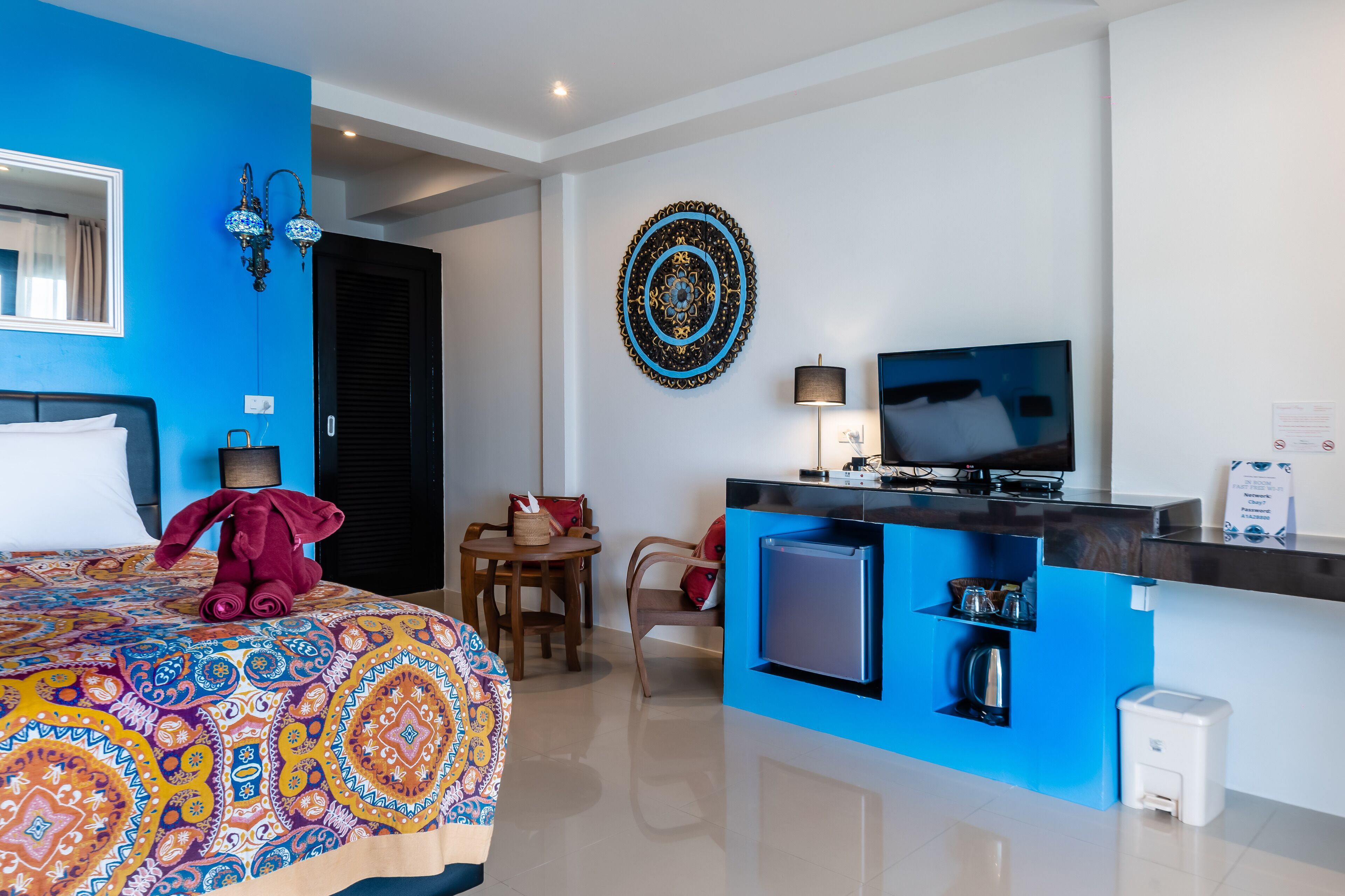 A hotel room with a bed, a desk with a TV, a small fridge, and a decorative wall piece.