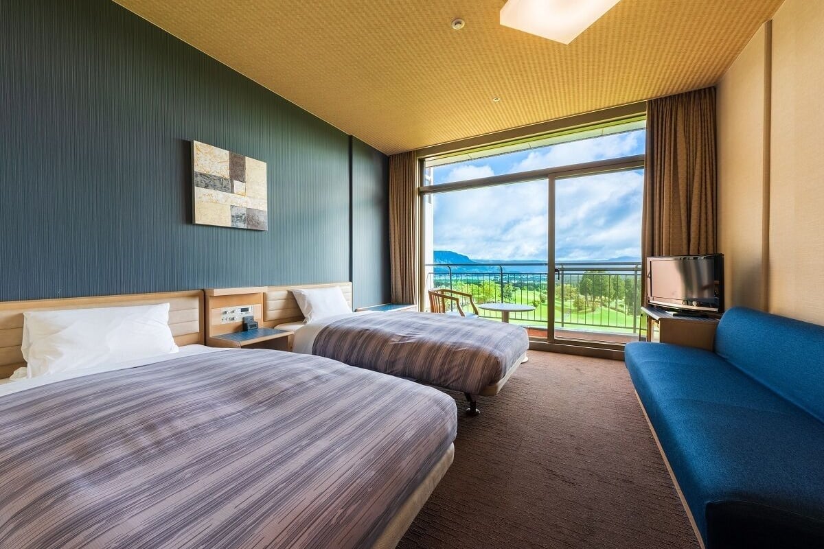 A hotel room with two beds, a sofa, a TV, and a balcony with a view of the sea.