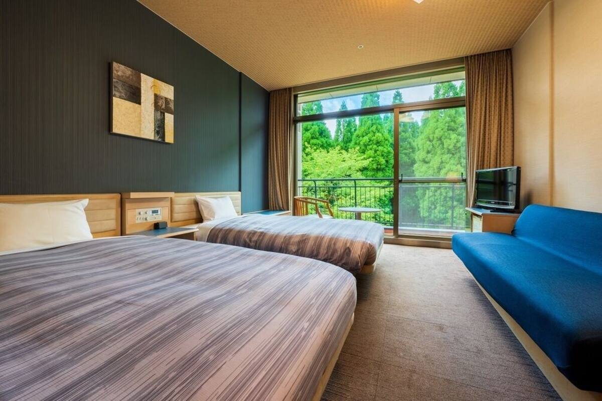 A hotel room with two beds, a sofa, a TV, and a balcony with a view of trees.