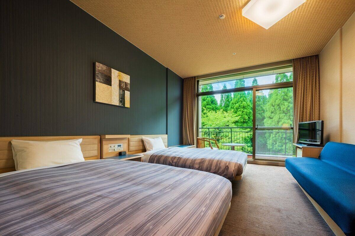 A hotel room with two beds, a sofa, a TV, and a balcony with a view of greenery.